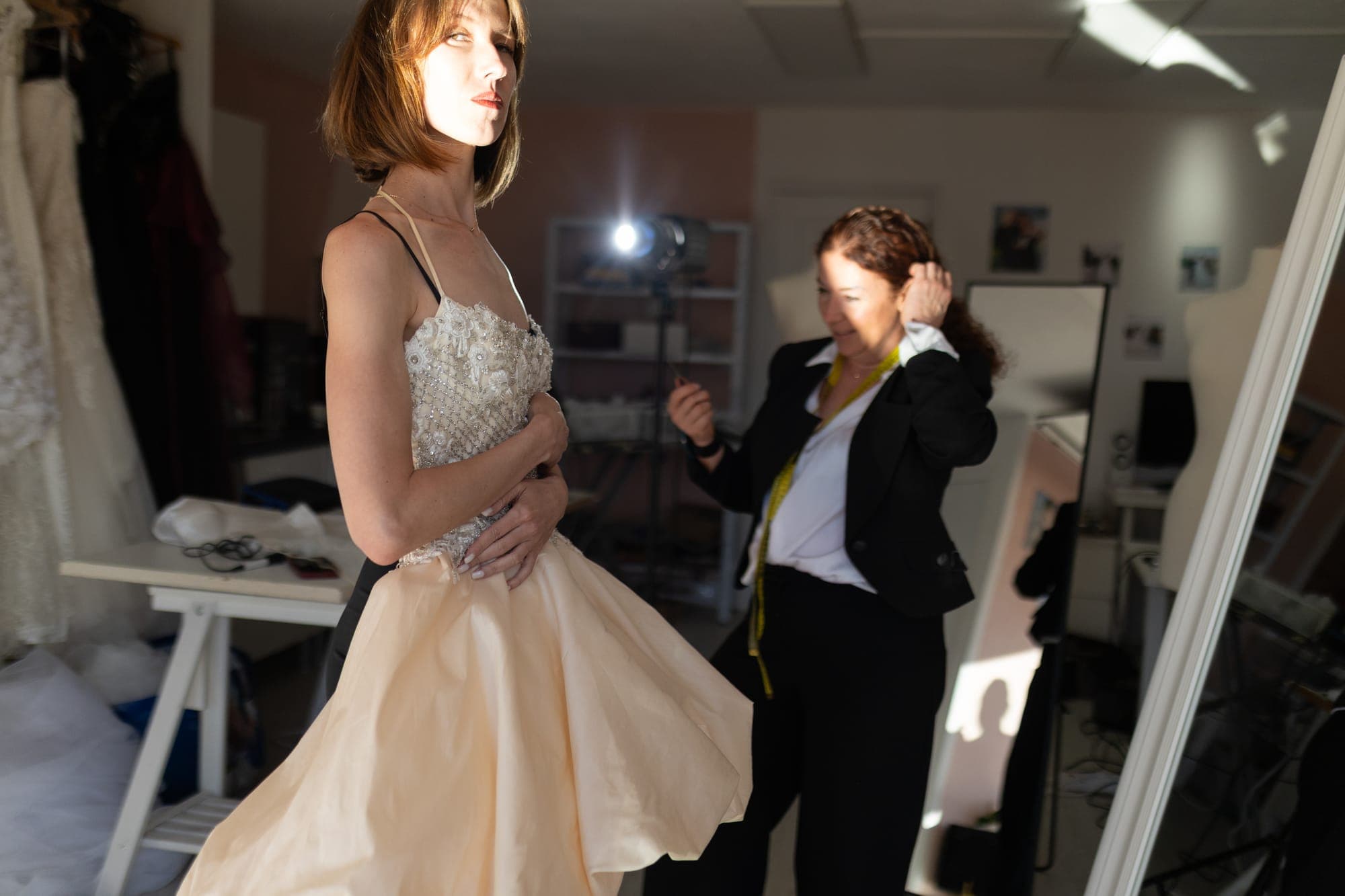 Model in a short champagne dress with a beaded bodice and full skirt, the designer adjusting it behind her