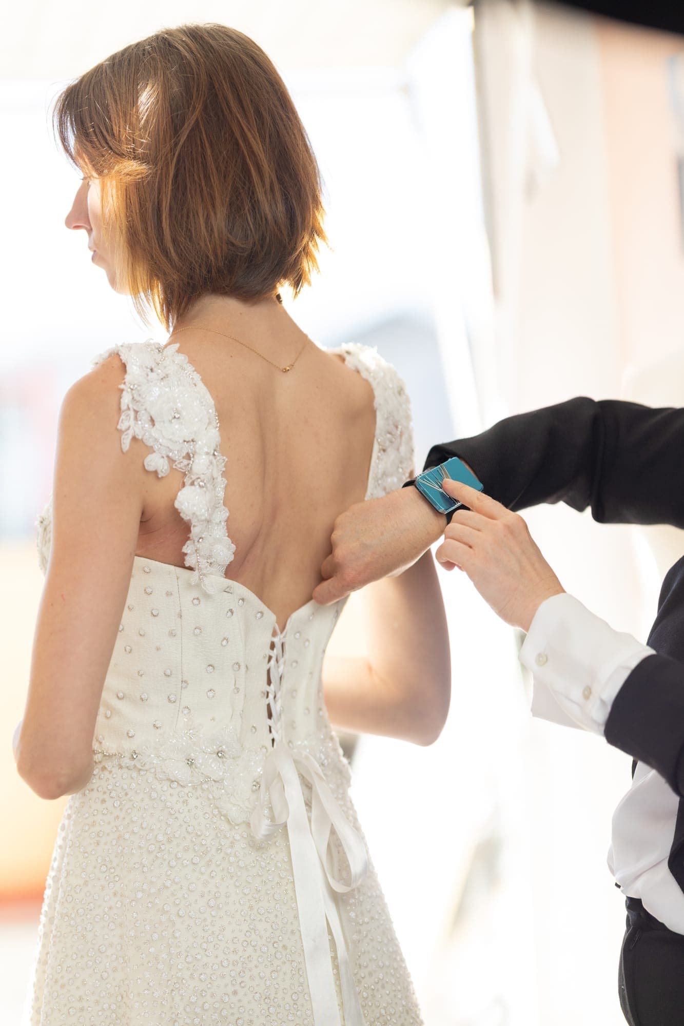 Stylist closing the laced corset back of a lace wedding gown on a bride during a fitting