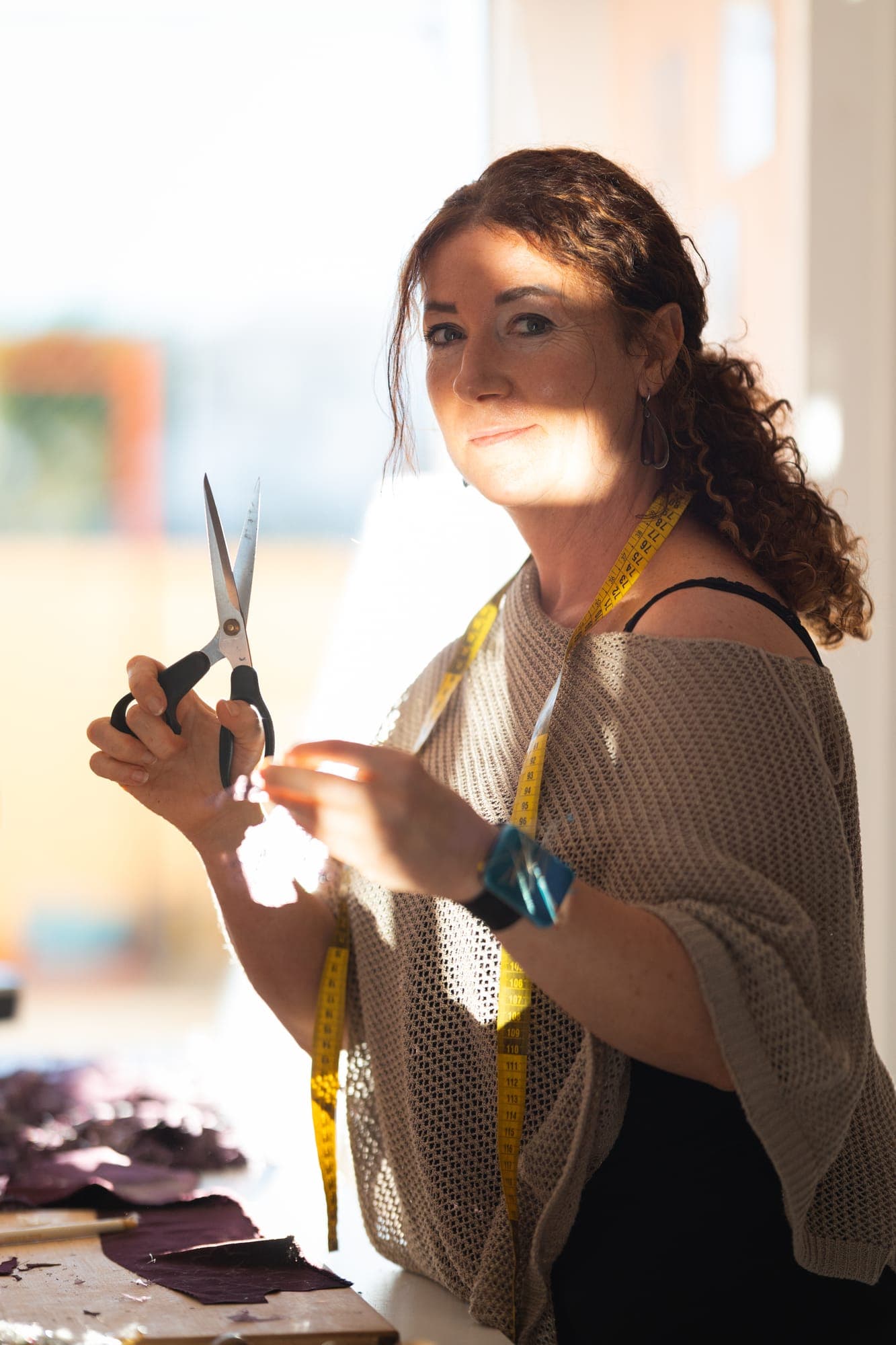 Smiling couturière holding tailor's shears and thread in a bright bridal atelier, a yellow measuring tape around her neck