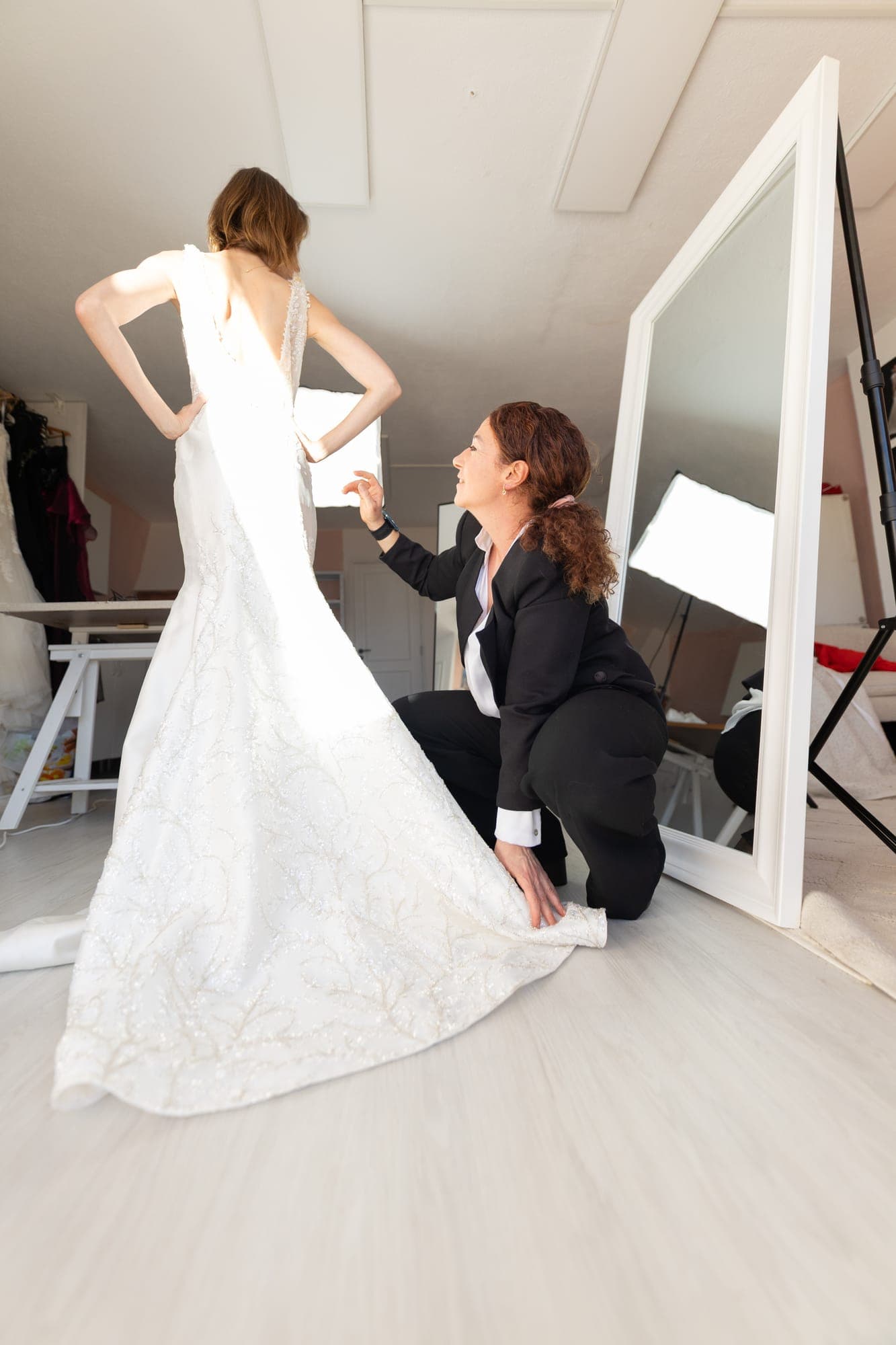 Designer kneeling to make a final hem adjustment to a fishtail lace wedding gown in front of a mirror