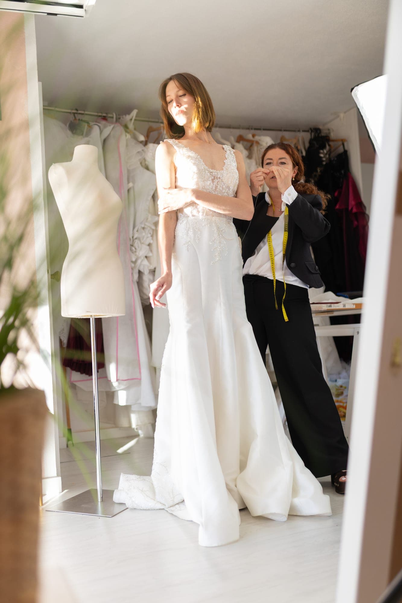 Bride standing in a lace wedding gown beside a dress form while the designer assesses the fit