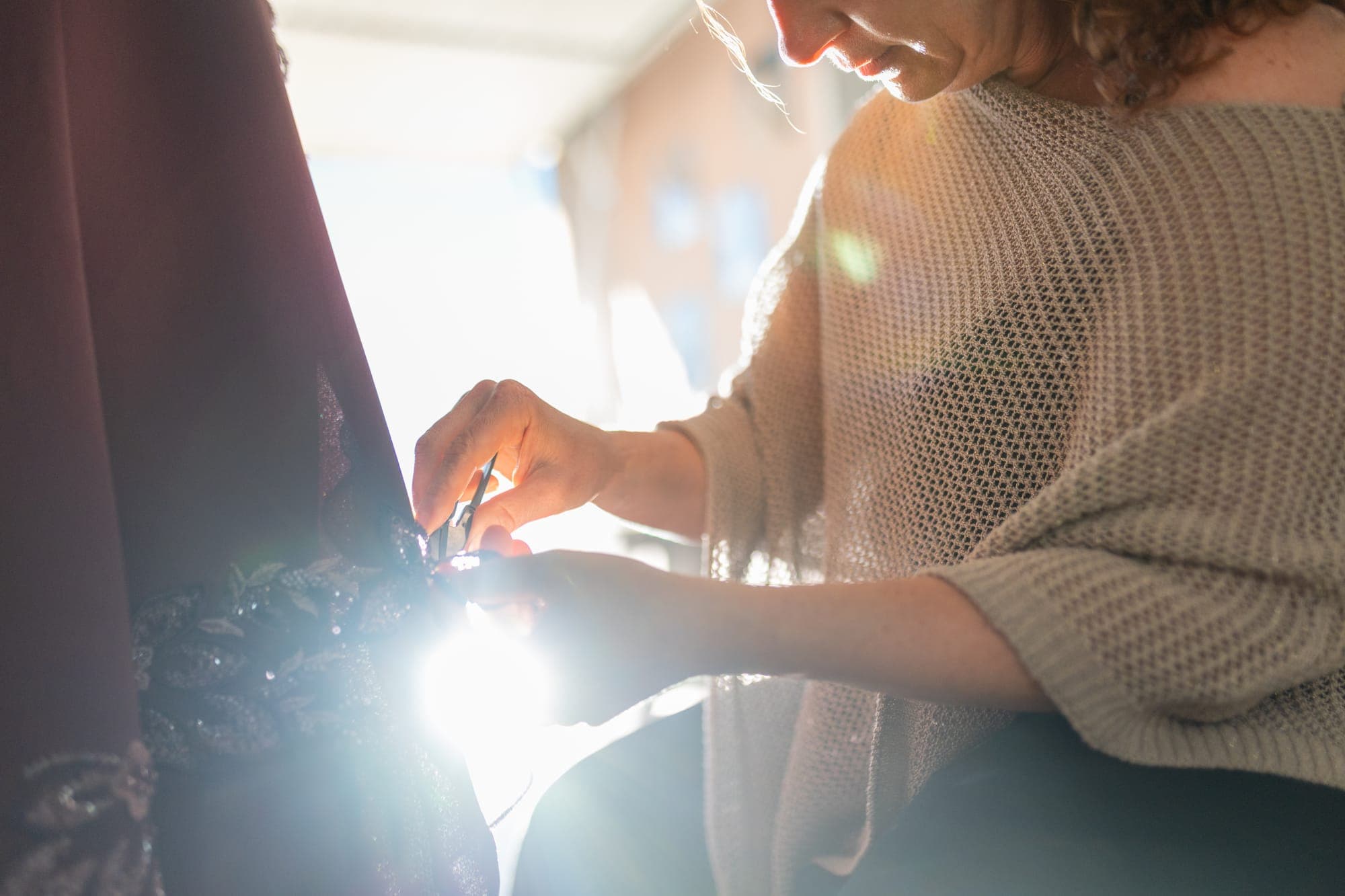 Couturière hand-stitching a beaded panel at an atelier workbench, backlit by strong window light