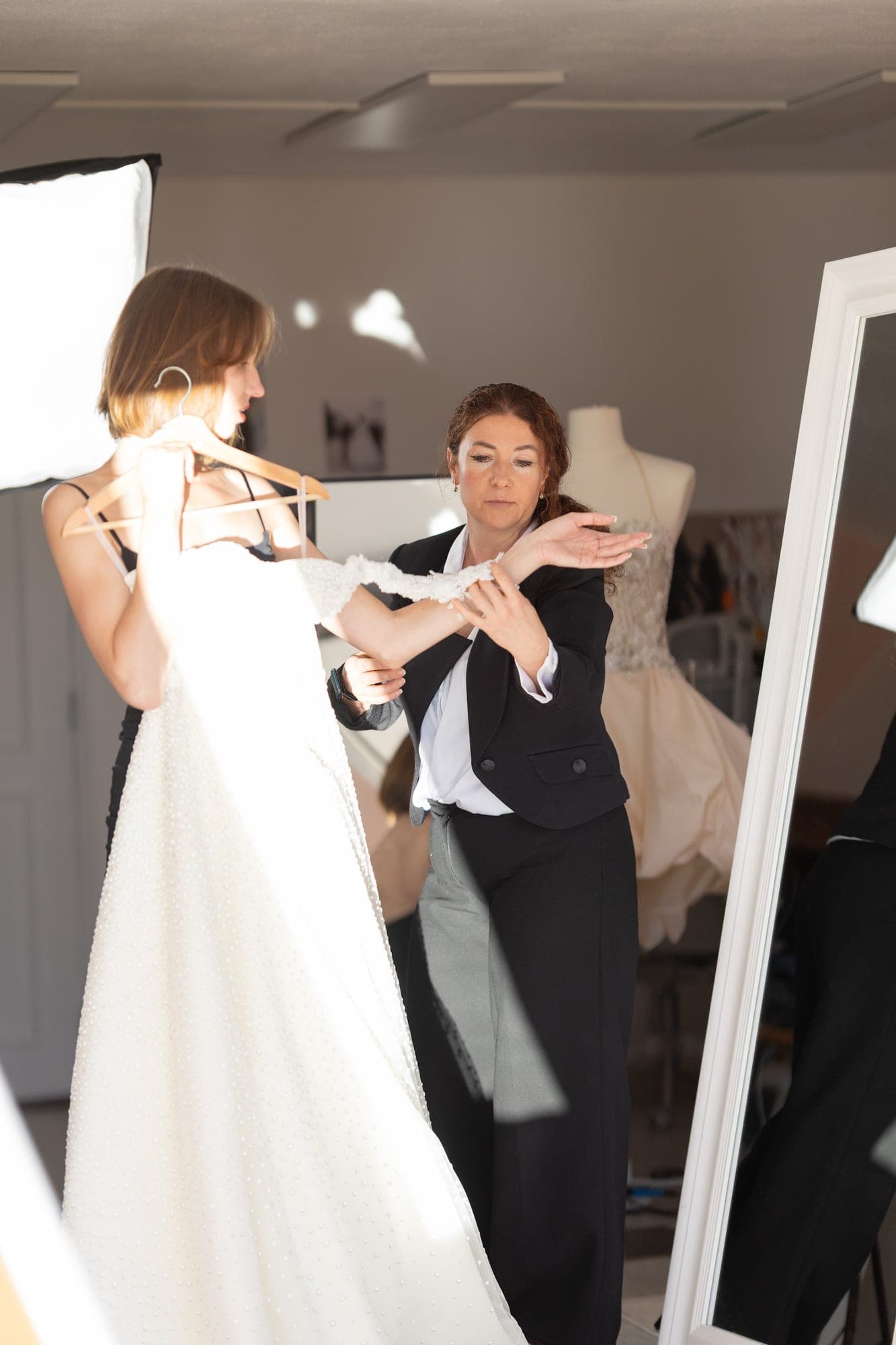 Fitter pinning the back of a lace wedding gown on a bride standing at a full-length mirror